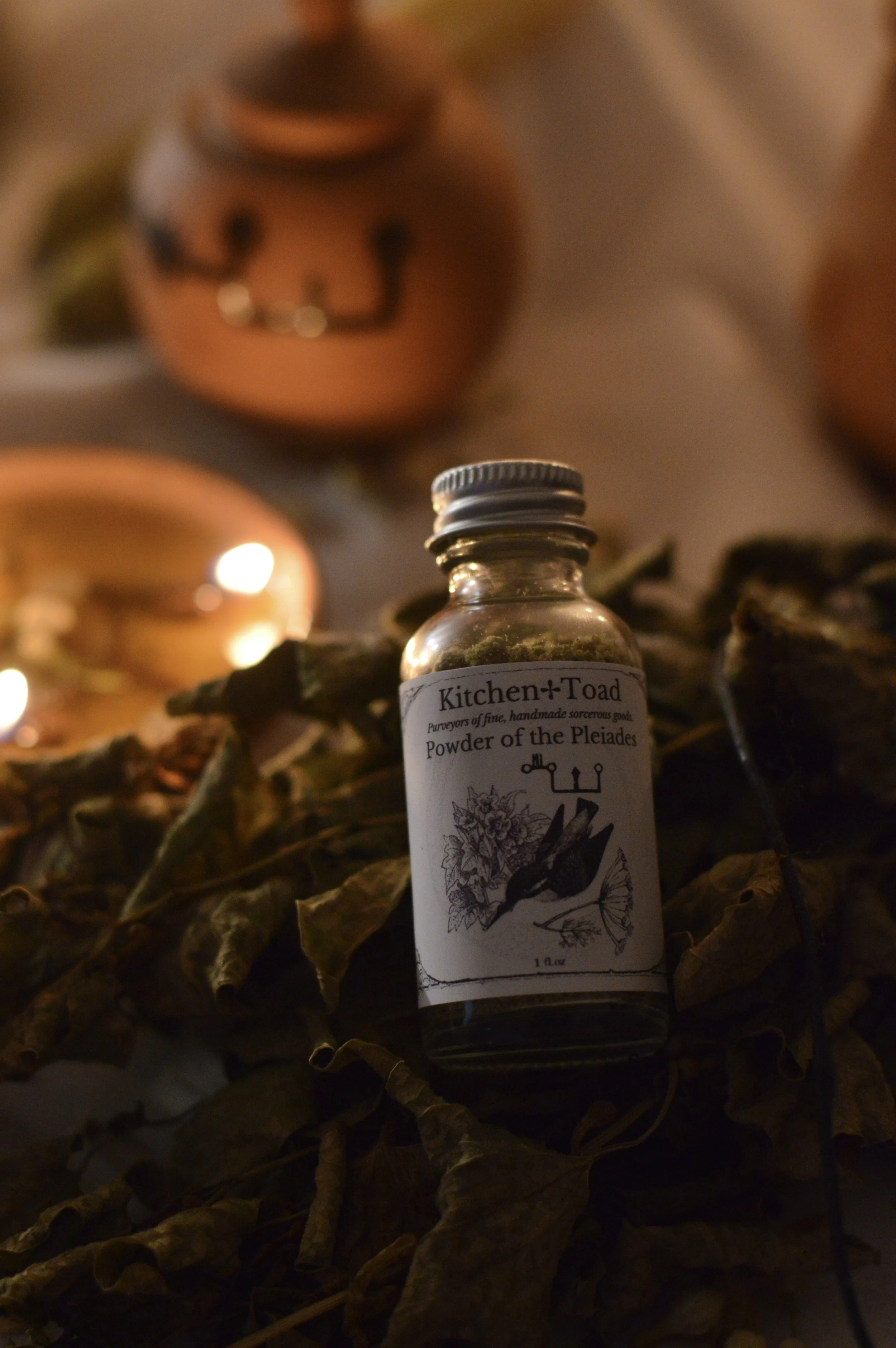 A small glass bottle labeled 'Kitchen Toad Powder of the Pleiades' rests on dried leaves, with a Halloween pumpkin decoration in the background.