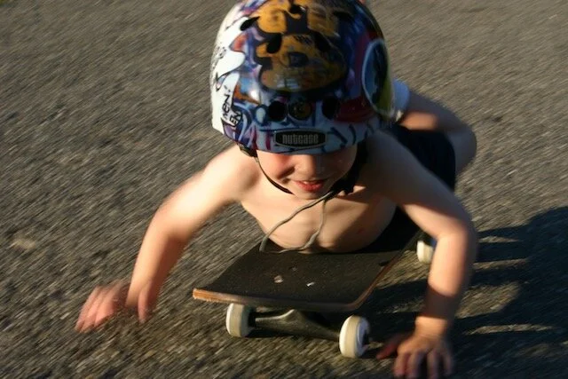 A young boy wearing a colorful helmet rides a skateboard on pavement.