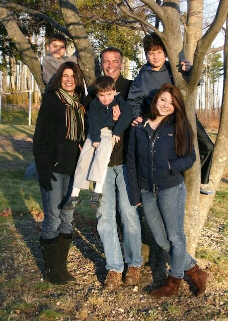 A family of six posing outdoors next to a tree, smiling. The group includes a man, three women, and two boys, all standing on grass with bare trees in the background.