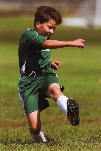 Young boy in green sports uniform kicking a soccer ball on a grassy field during daytime.