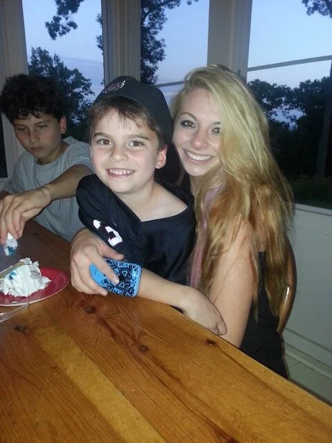 A group of three young people sitting around a wooden table indoors, with a woman embracing a smiling boy in the center, and another boy sitting nearby, with a window showing trees and sky in the background.