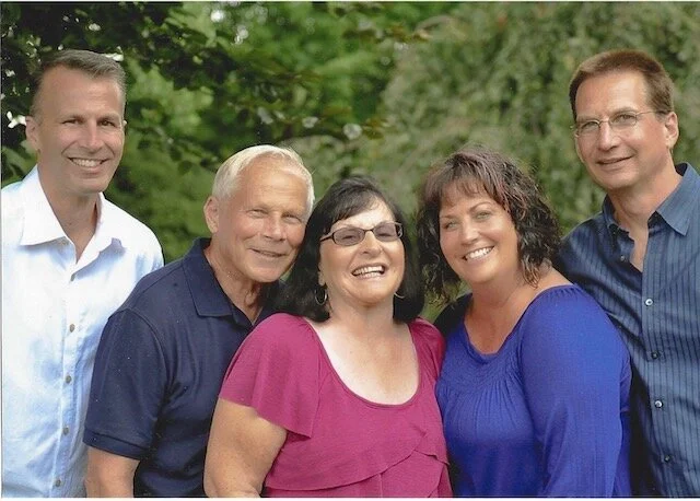 Group of five smiling adults outdoors in front of green trees