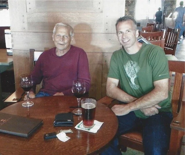 Two men sitting at a restaurant table with drinks, one older man with a beard and glasses and one younger man smiling, with wine glasses and a pint glass on the table.