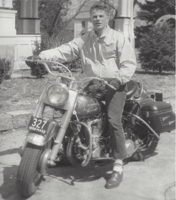 Young man sitting on a vintage motorcycle outdoors in front of a house with trees and bushes.