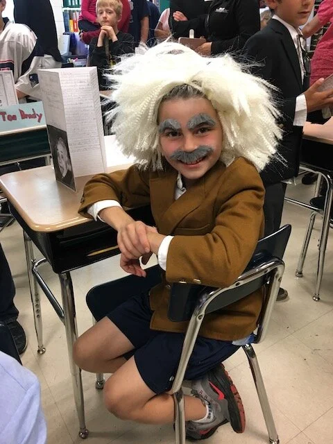 Child dressed as Albert Einstein with wild white wig, gray eyebrows and mustache, younger age. Sitting at school desk in classroom, smiling.