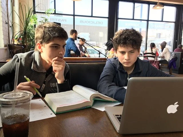 Two boys sitting at a table in a cafe, working on their laptops with notebooks open, while a musician plays guitar in the background.