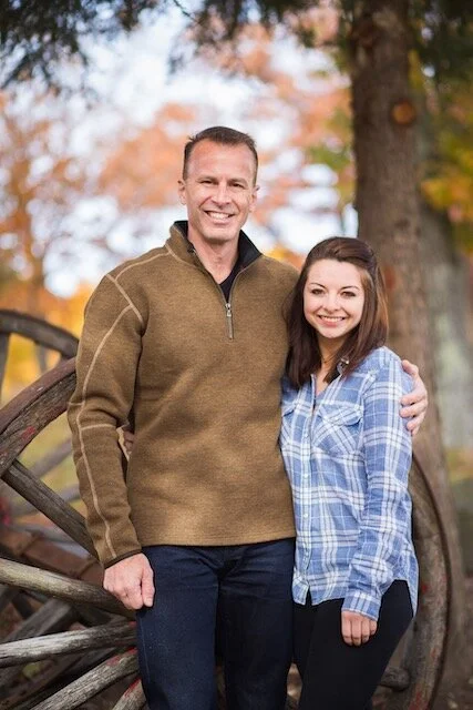 A man and a woman standing outdoors in front of a rustic wooden fence and trees with autumn leaves, smiling at the camera.