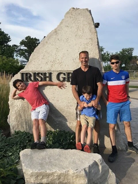 A family of four posing in front of a large rock with the words 'Irish Green' engraved on it, outdoors on a sunny day.