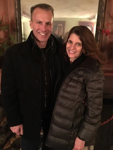 A man and woman smiling and standing together indoors, with the man wearing a black coat and the woman wearing a dark puffer jacket.