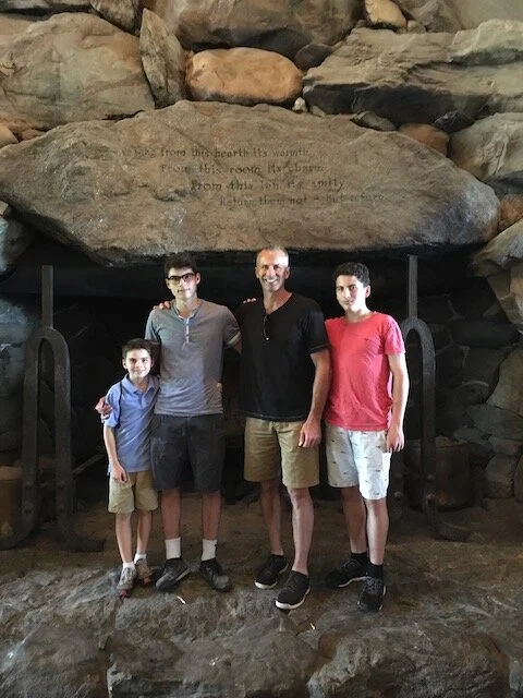 Four people standing in front of a large stone with an inscription, inside a cave or rocky area.