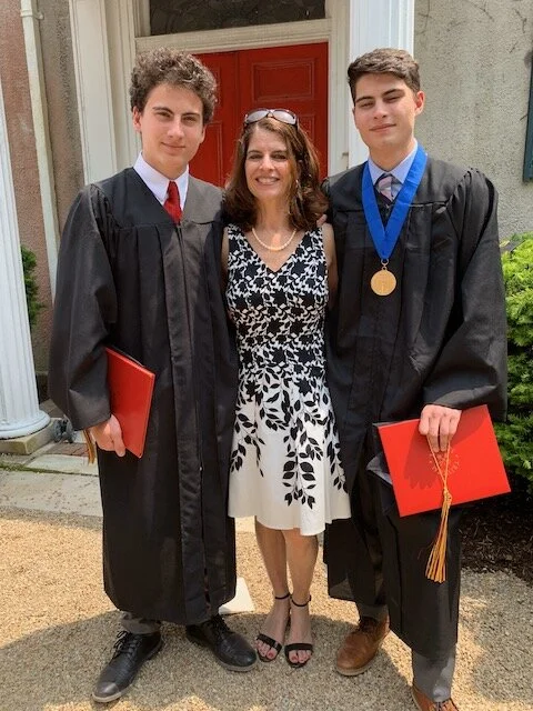 Two young men in graduation caps and gowns standing on either side of a woman in a black and white dress, in front of a red door, with one holding a red diploma folder and the other holding a red diploma folder and a medal around his neck.