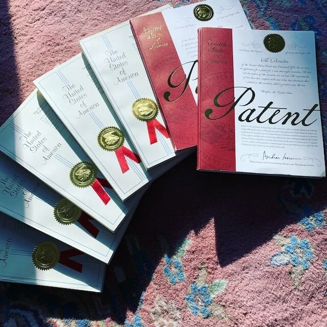 A fan of multiple patents laid out on a pink and blue floral carpet, with the front book titled 'Patent' and others titled 'The United States of America' with gold seals.
