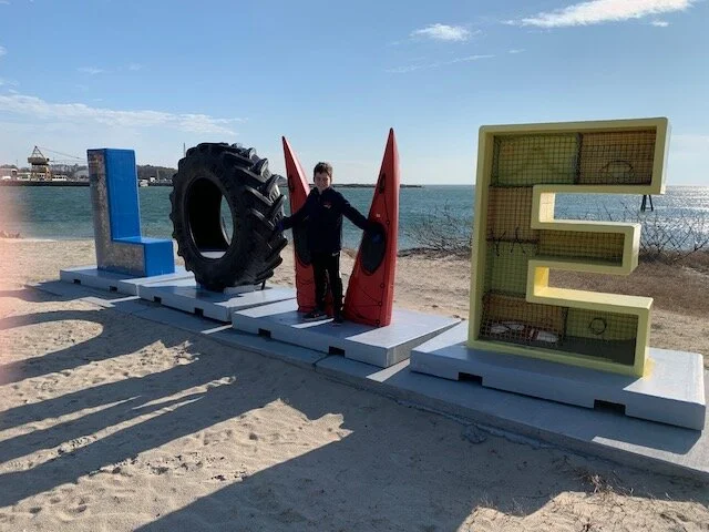 Person standing behind large, colorful letters spelling 'LOVE' on a beach, with the 'O' replaced by a tire, the 'V' by two red surfboards, and the 'E' by a yellow and green decorative structure.