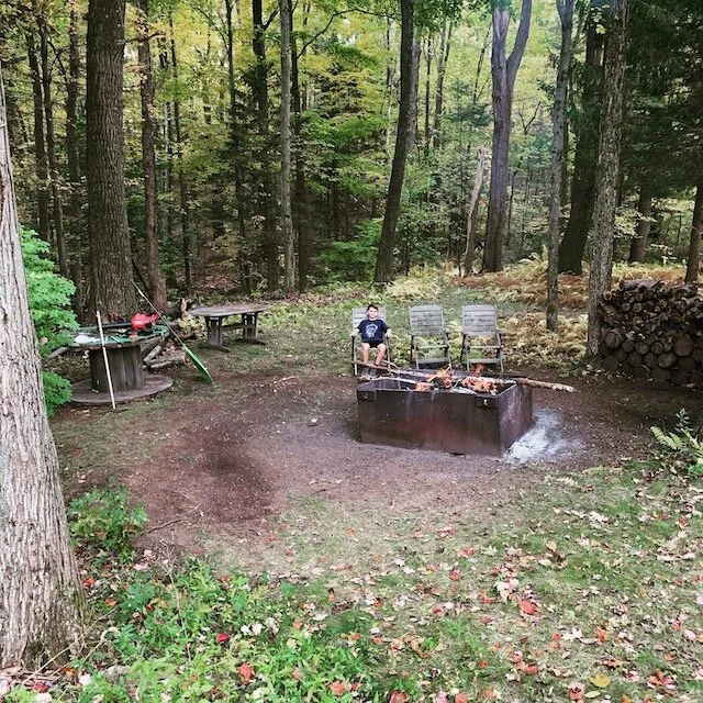 Child sitting on a chair near a fire pit in a wooded outdoor area with trees and logs.