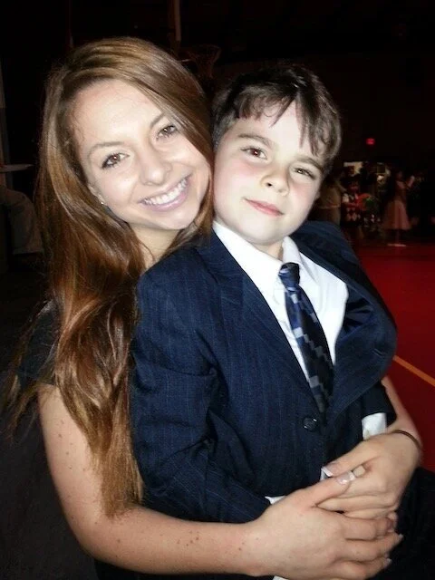 A woman with long reddish-brown hair and a boy with short brown hair, both smiling, dressed in formal attire, posing together at an indoor event.