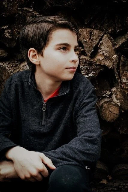 Young boy with dark hair in profile sitting outdoors against a background of stacked logs, wearing a dark zip-up jacket.