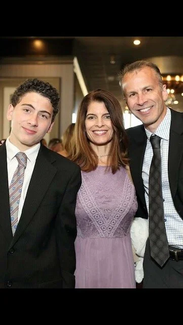 A family of three smiling at the camera, dressed in formal attire, indoors at a celebratory event.