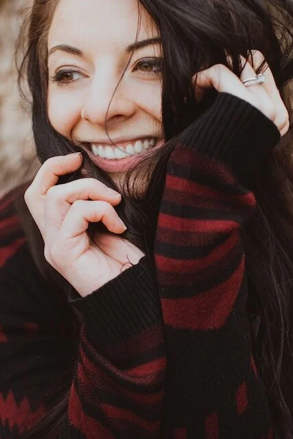 Close-up of a smiling woman with long dark hair, wearing a black and red striped sweater, with her hand near her face.