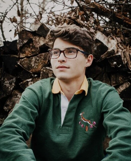 Young man with glasses wearing a green and yellow collared shirt sitting outdoors in front of a woodpile