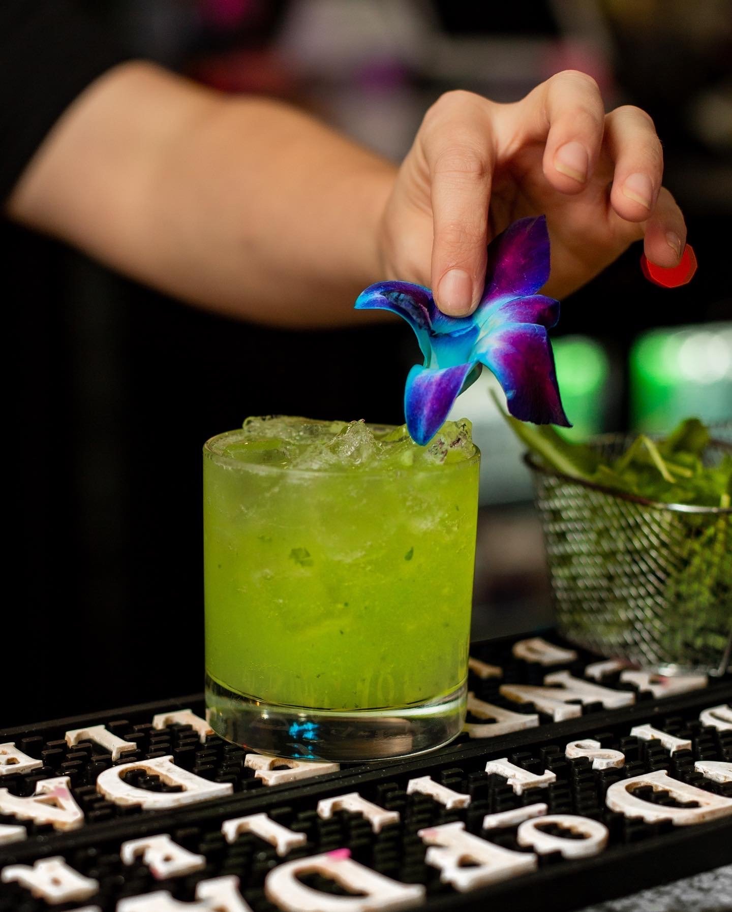 A hand placing a blue and purple flower on top of a green cocktail in a glass.