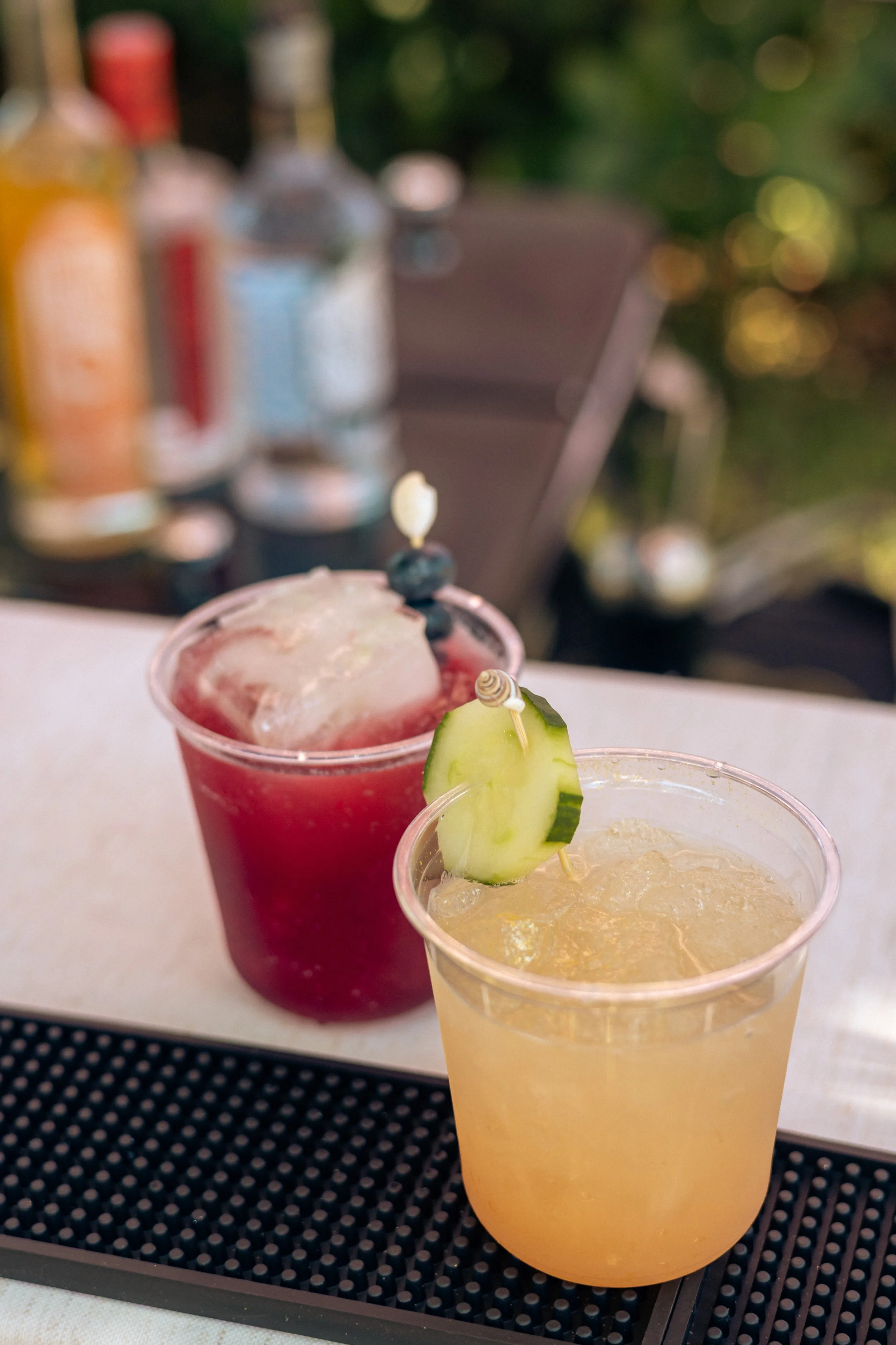 Two colorful cocktails on a bar or serving tray, one red and one yellow, garnished with a slice of cucumber, with blurred bottles and a chair in the background.