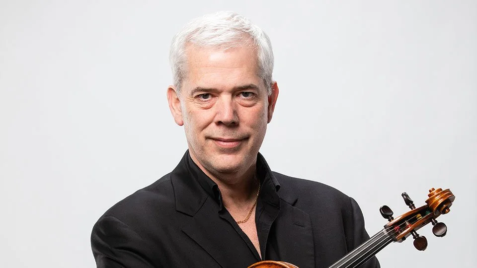 A man with gray hair wearing a tuxedo and bow tie, holding a violin.