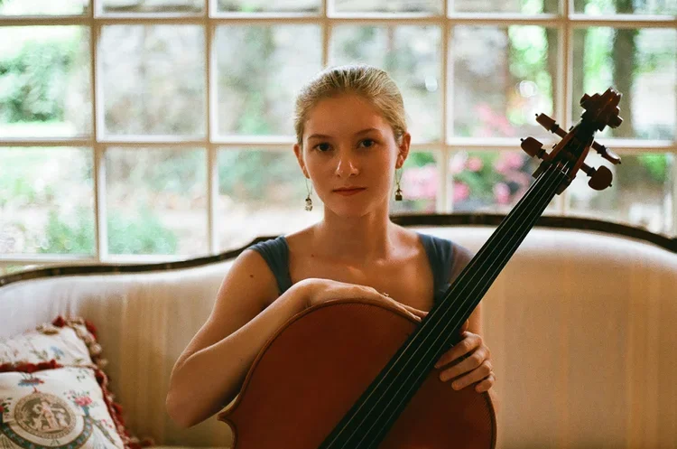 A woman with long brown hair, wearing a black sheer top, sits behind a large wooden cello with both arms resting on it, against a dark background.