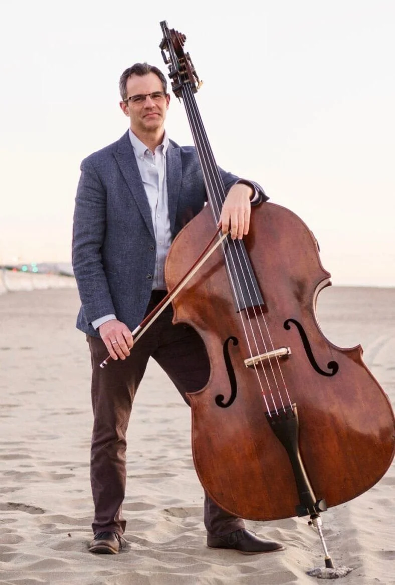 A smiling woman with long wavy brown hair wearing a black lace dress, holding a bow and a cello.
