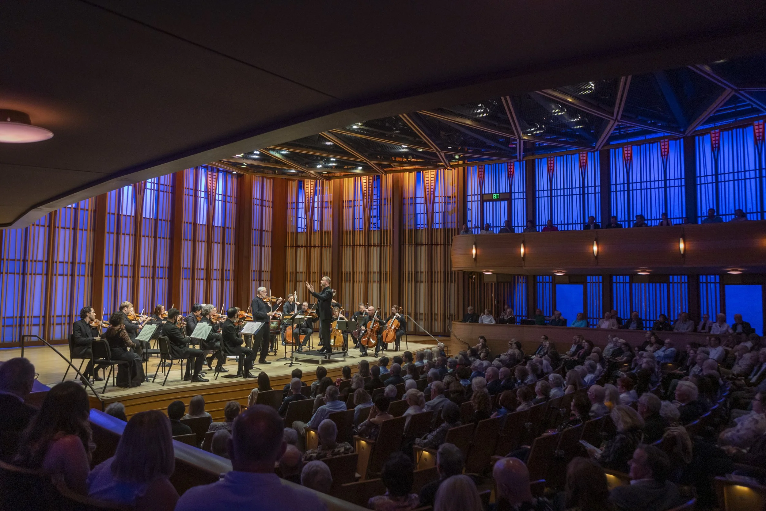 A conductor leading an orchestra during a performance in a concert hall with audience members watching.