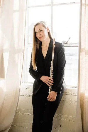 A young woman with long brown hair wearing a brown leather jacket, holding a silver flute, standing outdoors by a concrete wall in an urban setting.