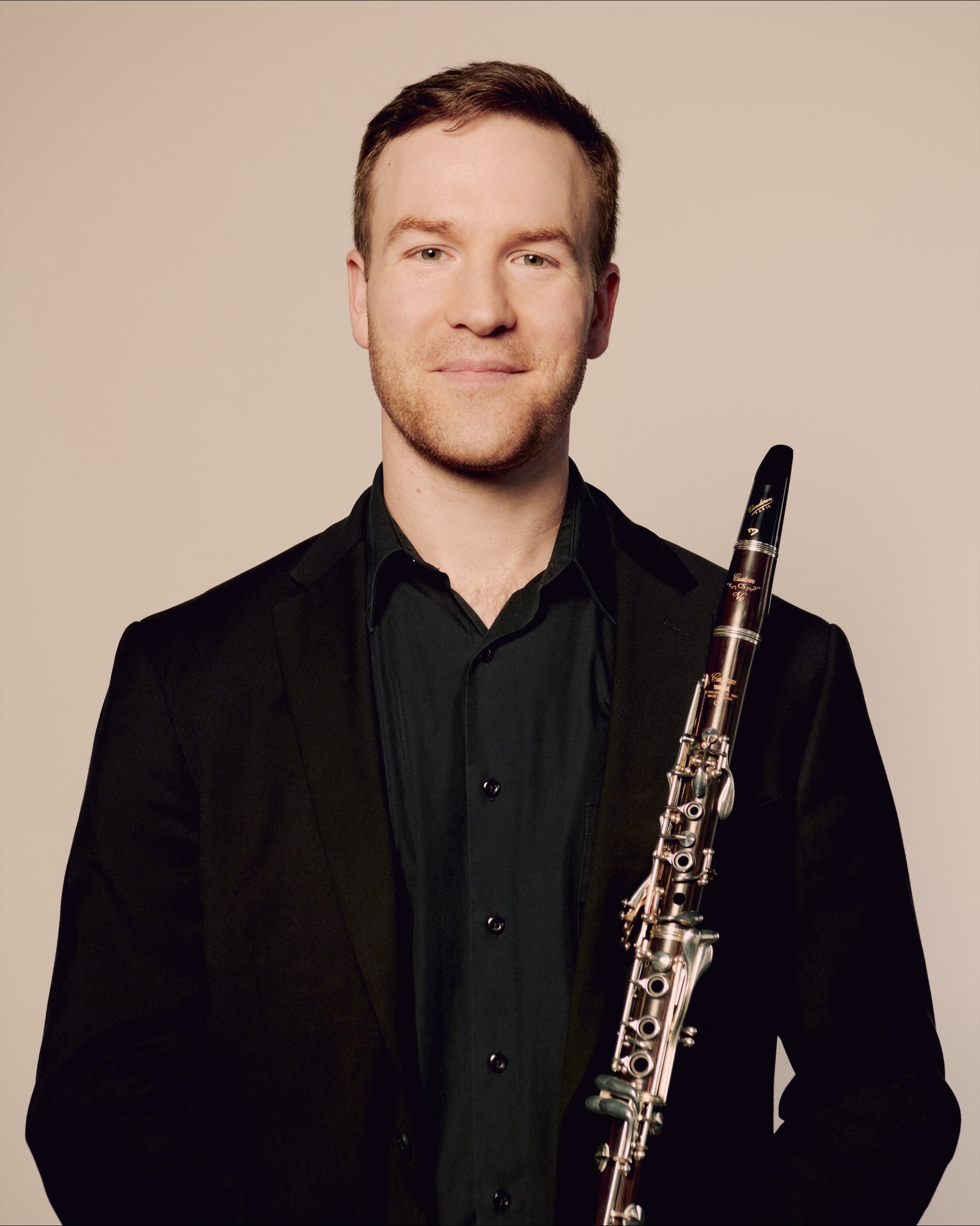 A man with a beard, wearing a black shirt and suit jacket, holding a clarinet, smiling at the camera.