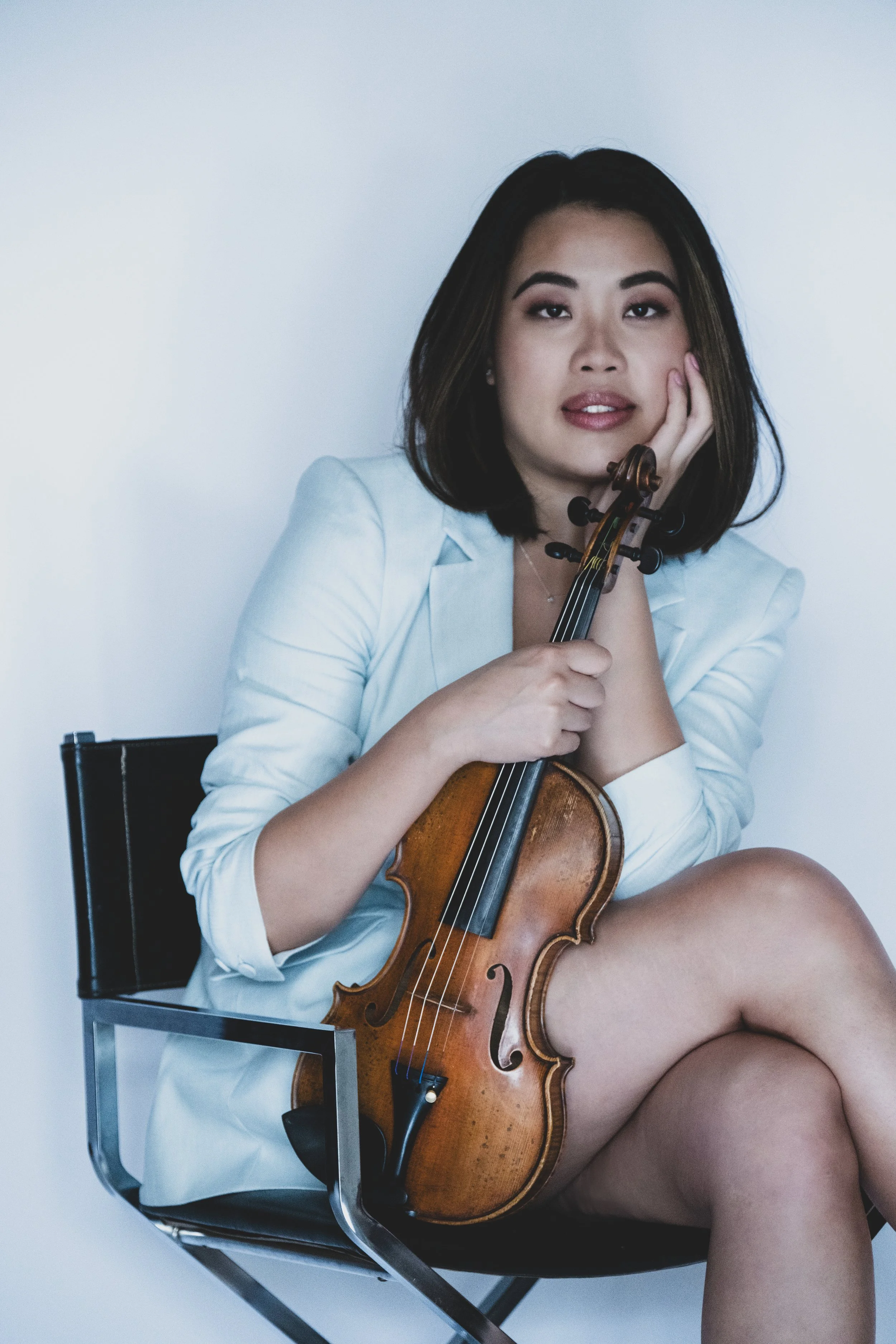 A young woman with long dark hair holding a violin, smiling softly. She is wearing a light-colored top and is standing in a well-lit outdoor area with blurred wooden steps in the background.
