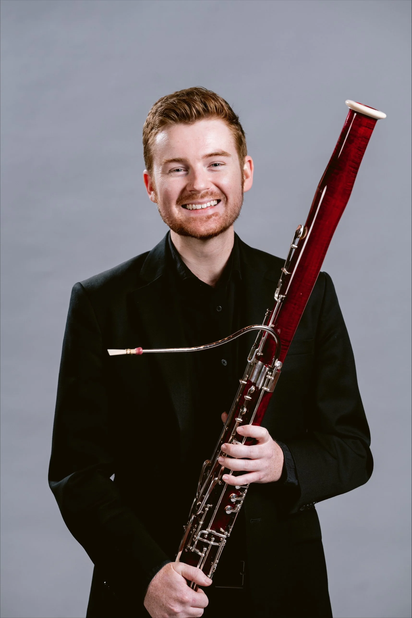 A young man with red hair, glasses, and a light blue shirt smiling while holding a large red bassoon.