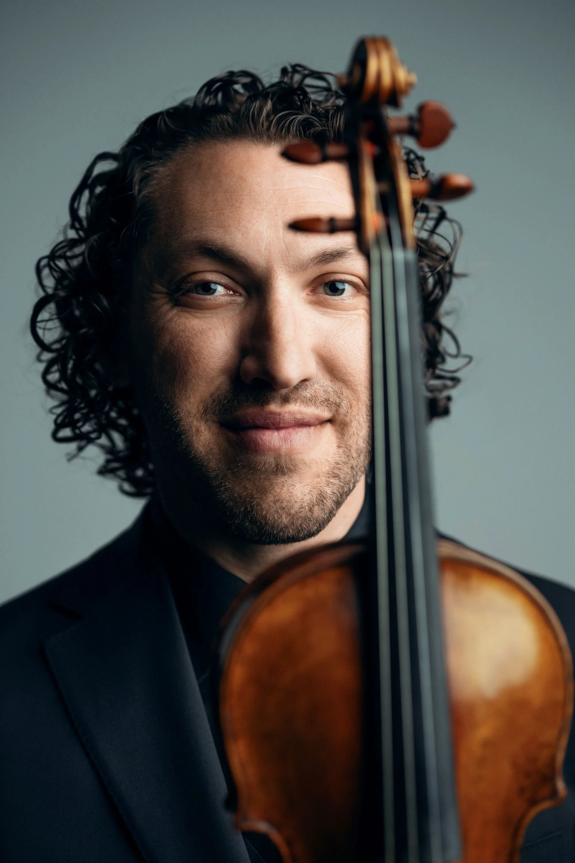 Man in black suit holding a violin, standing outdoors with a cityscape and marina in the background.