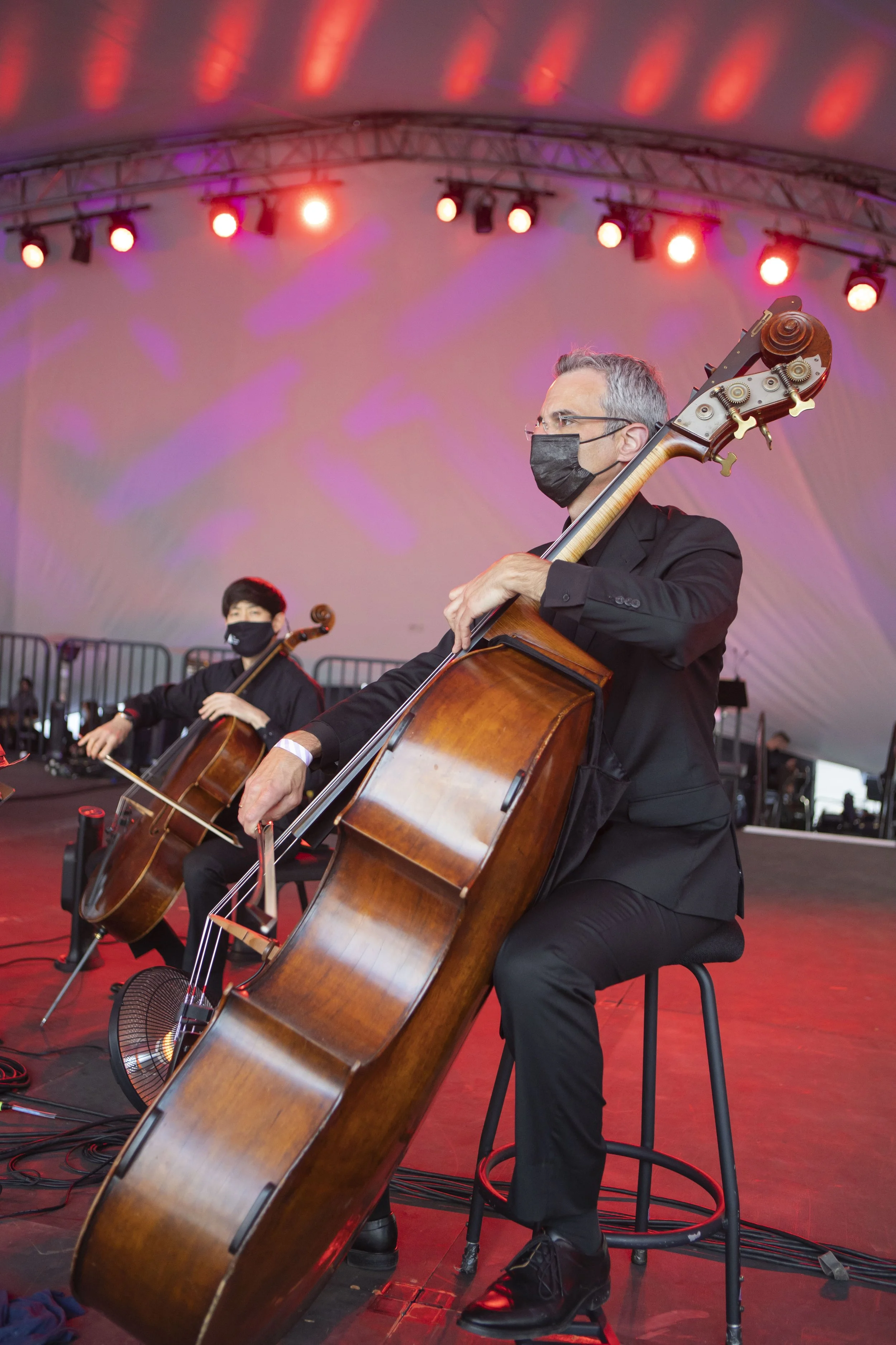 Two musicians wearing black suits and face masks playing cellos on stage with colorful lighting and a large white backdrop.