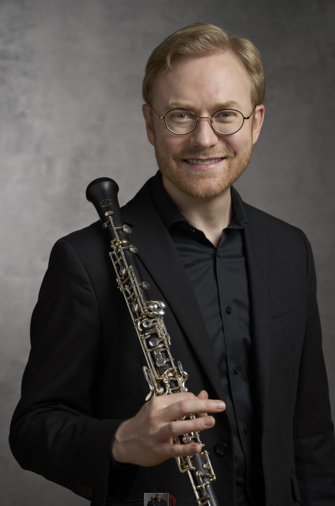 A man with glasses and a beard smiling, wearing a black suit and black shirt, holding a clarinet, against a plain white background.