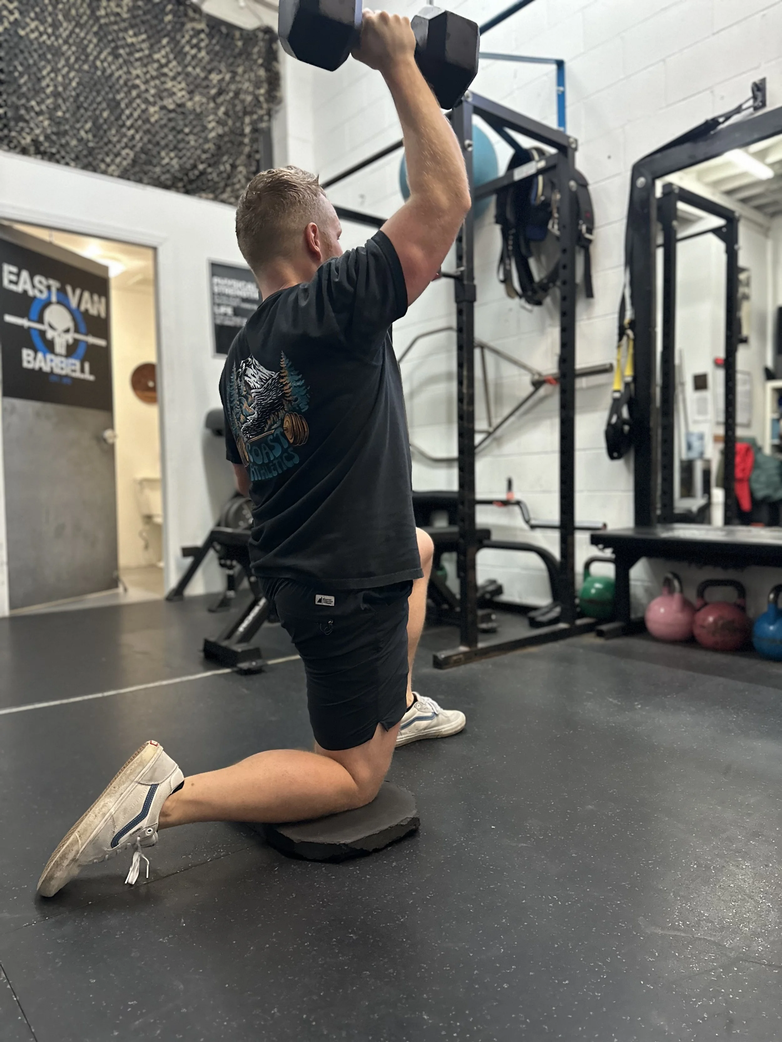 Man kneeling on one knee on a padded mat while lifting a dumbbell overhead in a gym.