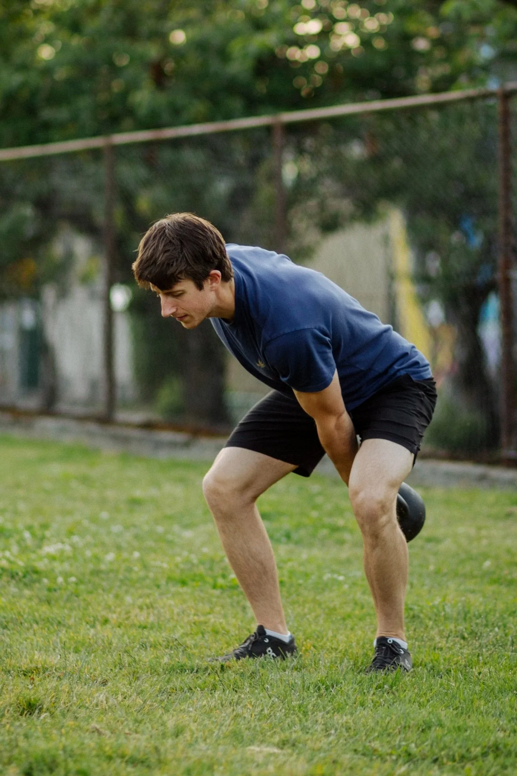Young man in athletic clothing performing a bent-over kettlebell swing exercise outdoors.