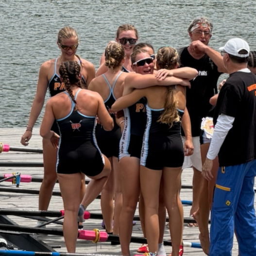 A group of women in rowing uniforms hugging and celebrating on a dock near the water.
