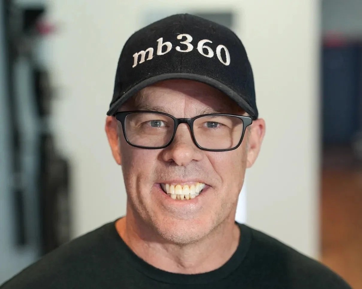 A man with glasses wearing a black baseball cap with 'mb360' embroidered on it, smiling at the camera.