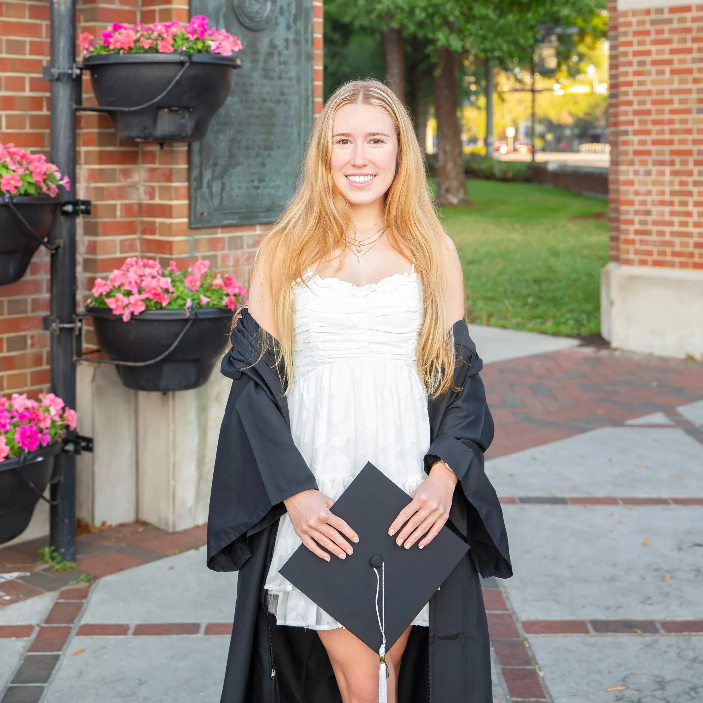 Teenage girl with long blonde hair smiling outdoors in front of a brick wall with flower planters, holding a black graduation cap in front of her waist.