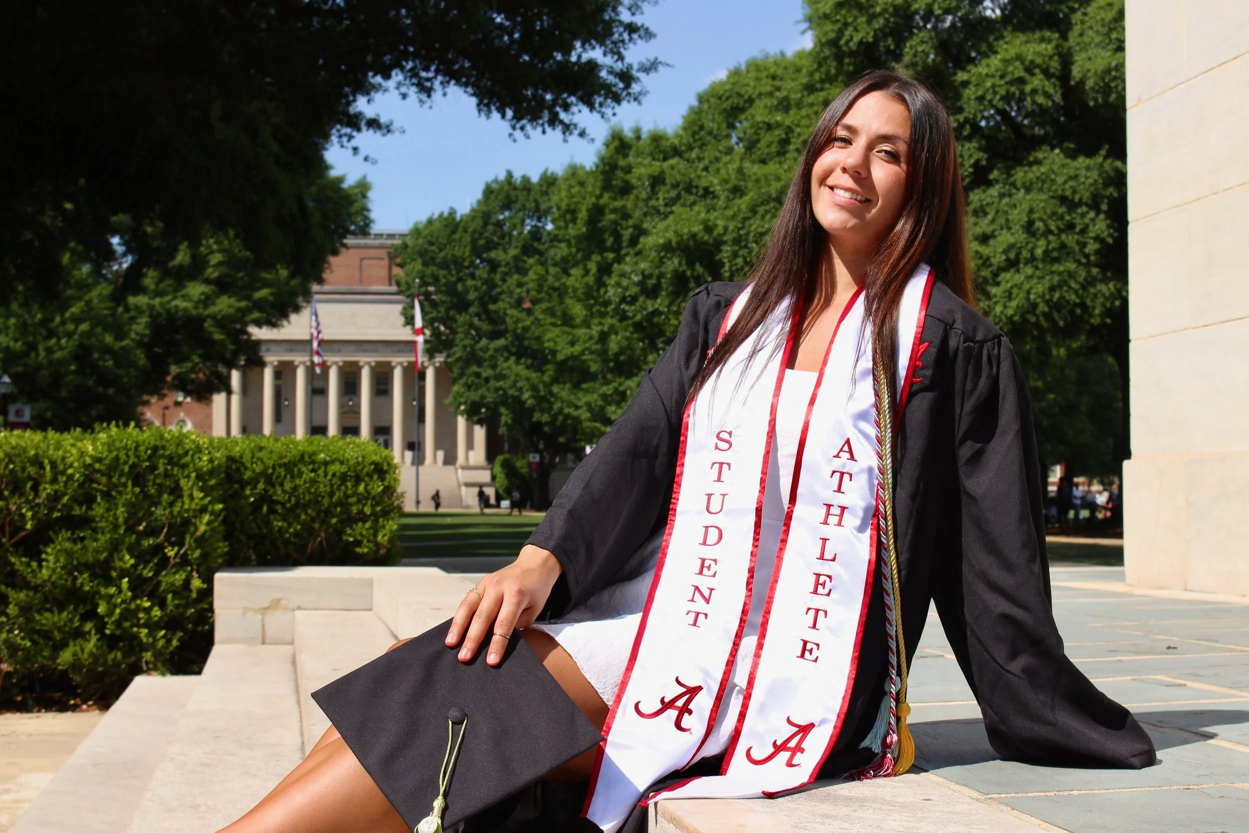 A young woman in a graduation cap and gown sitting outdoors on steps, smiling at the camera with a university building and trees in the background.