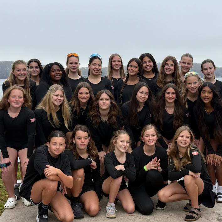 Group of young girls in black sports outfits outdoors by a body of water, posing for a team photo.