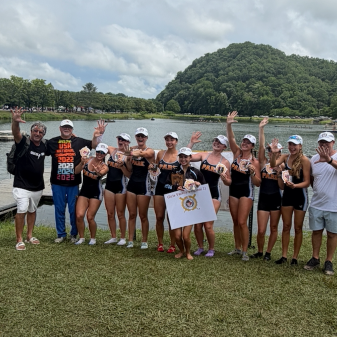 A group of young female athletes, along with their coaches, celebrating with medals and a flag near a lakeside with lush green trees and a mountain in the background.