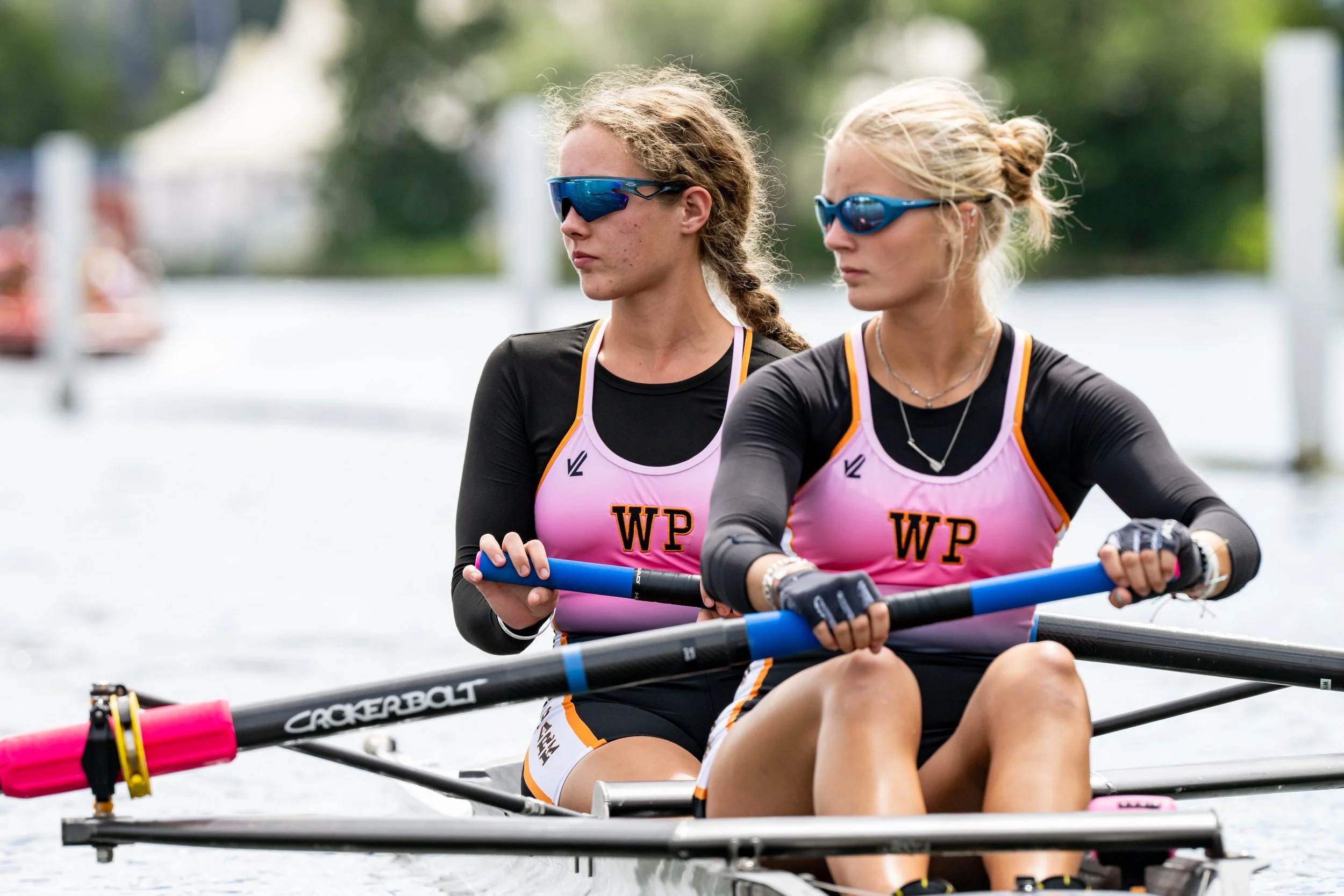 Two women rowing a boat on a river or lake, wearing sunglasses, black athletic clothing with pink jerseys that have 'WP' on them, and focused expressions.