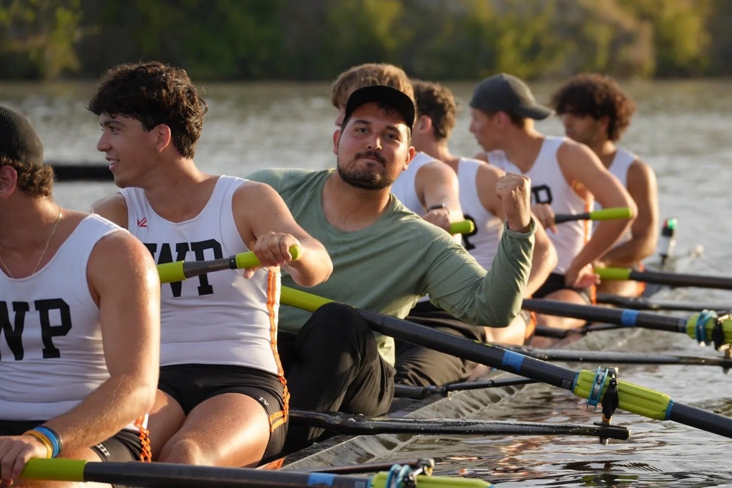 A team of rowers in a boat on a lake, wearing coordinated athletic gear, with the main rower flexing his arm and looking confidently at the camera.