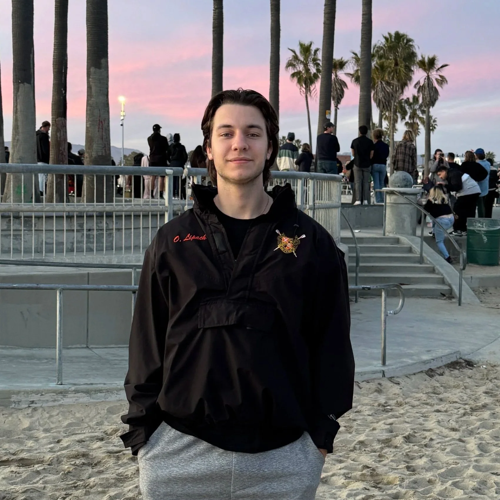 A young man with dark, shoulder-length hair standing on a beach at sunset, wearing a black jacket with emblem and light grey sweatpants, with palm trees and a crowd in the background.