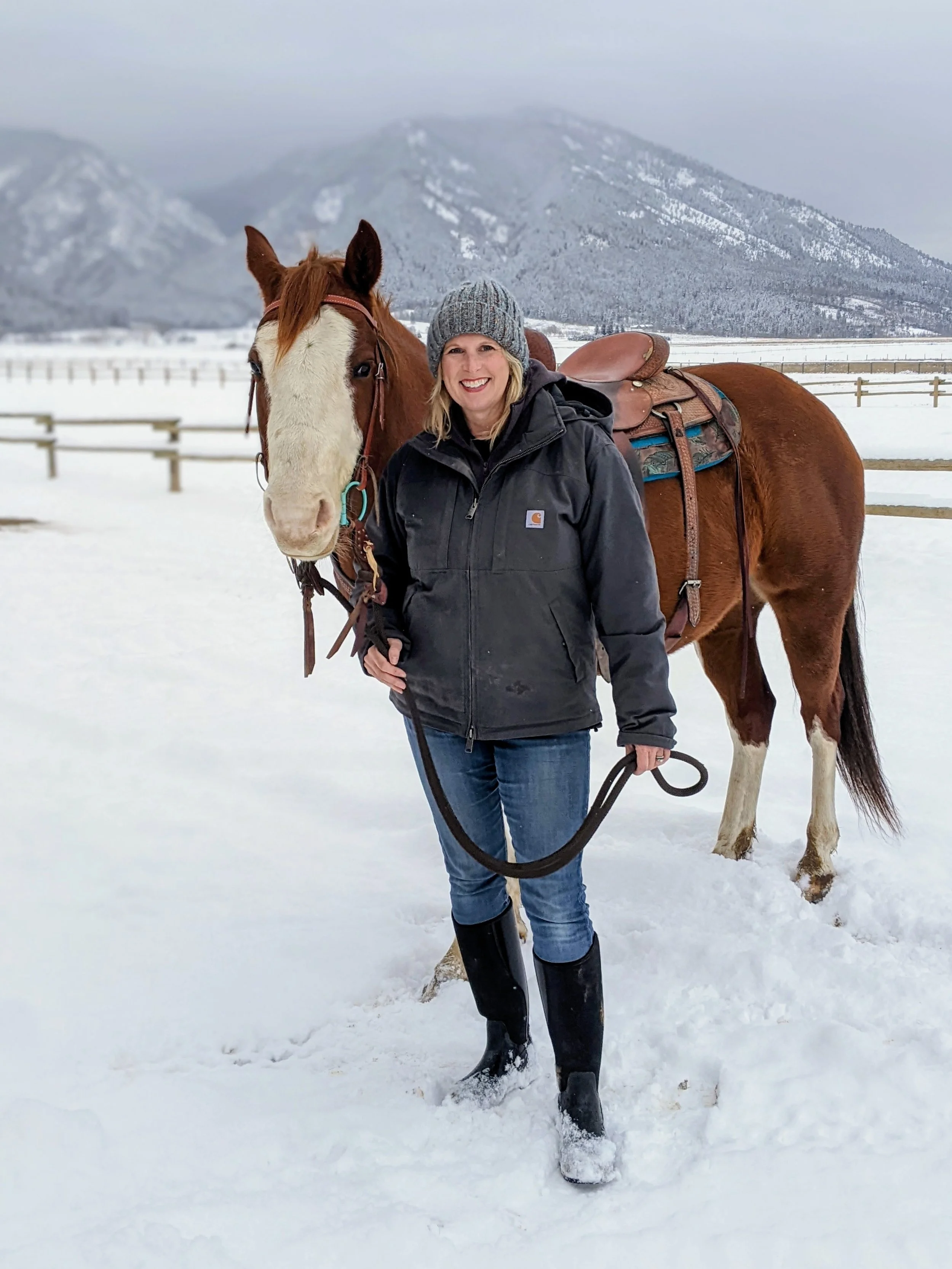 Woman in black jacket and gray beanie standing in snow with brown and white horse in a snowy field, mountains in the background.