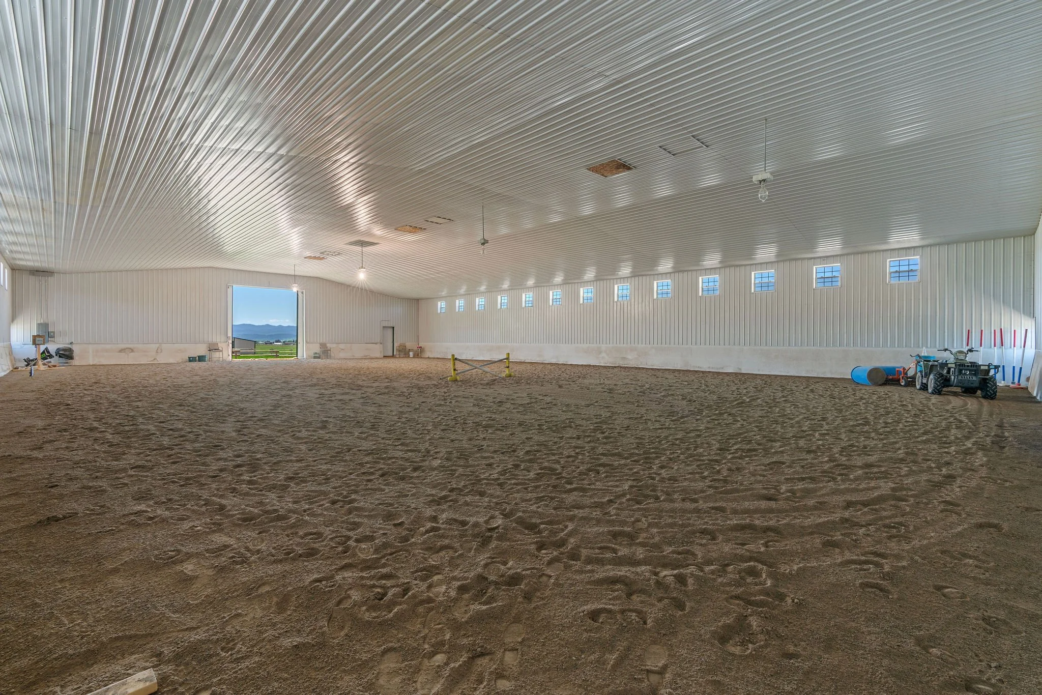 An indoor riding arena with dirt floor, white metal paneled walls, high ceiling with lights, small windows near the ceiling, outdoor landscape with mountains visible through an open doorway.