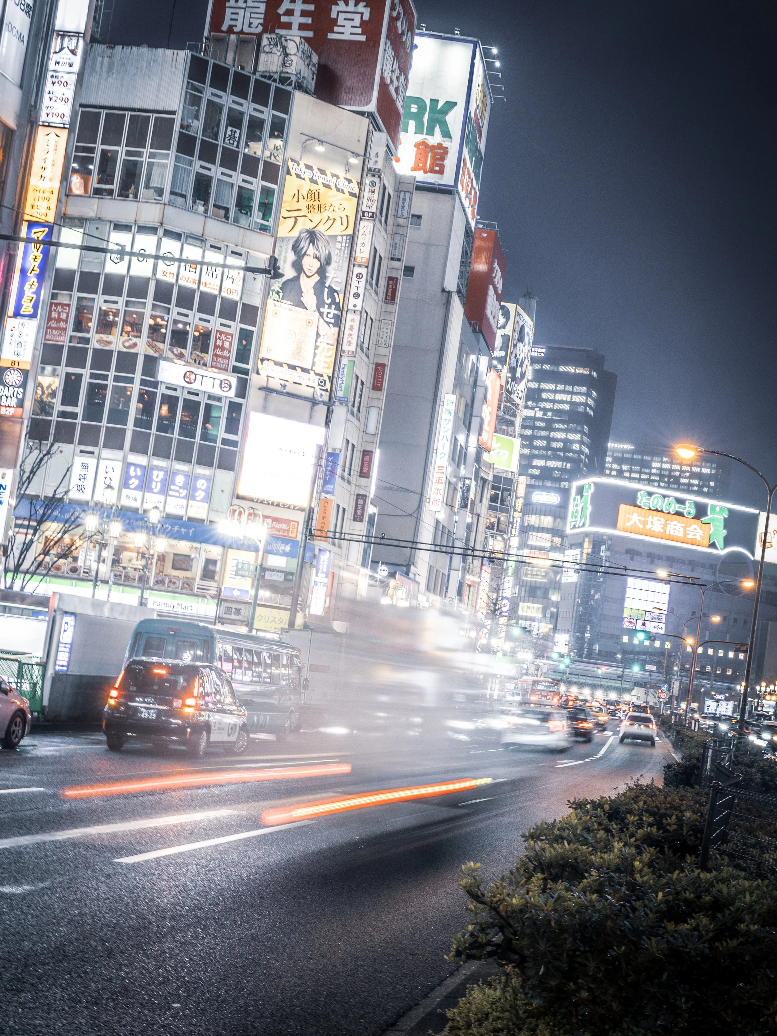 A bustling city street at night with bright neon signs and billboards in an urban area, featuring fast-moving cars and a mix of tall buildings.